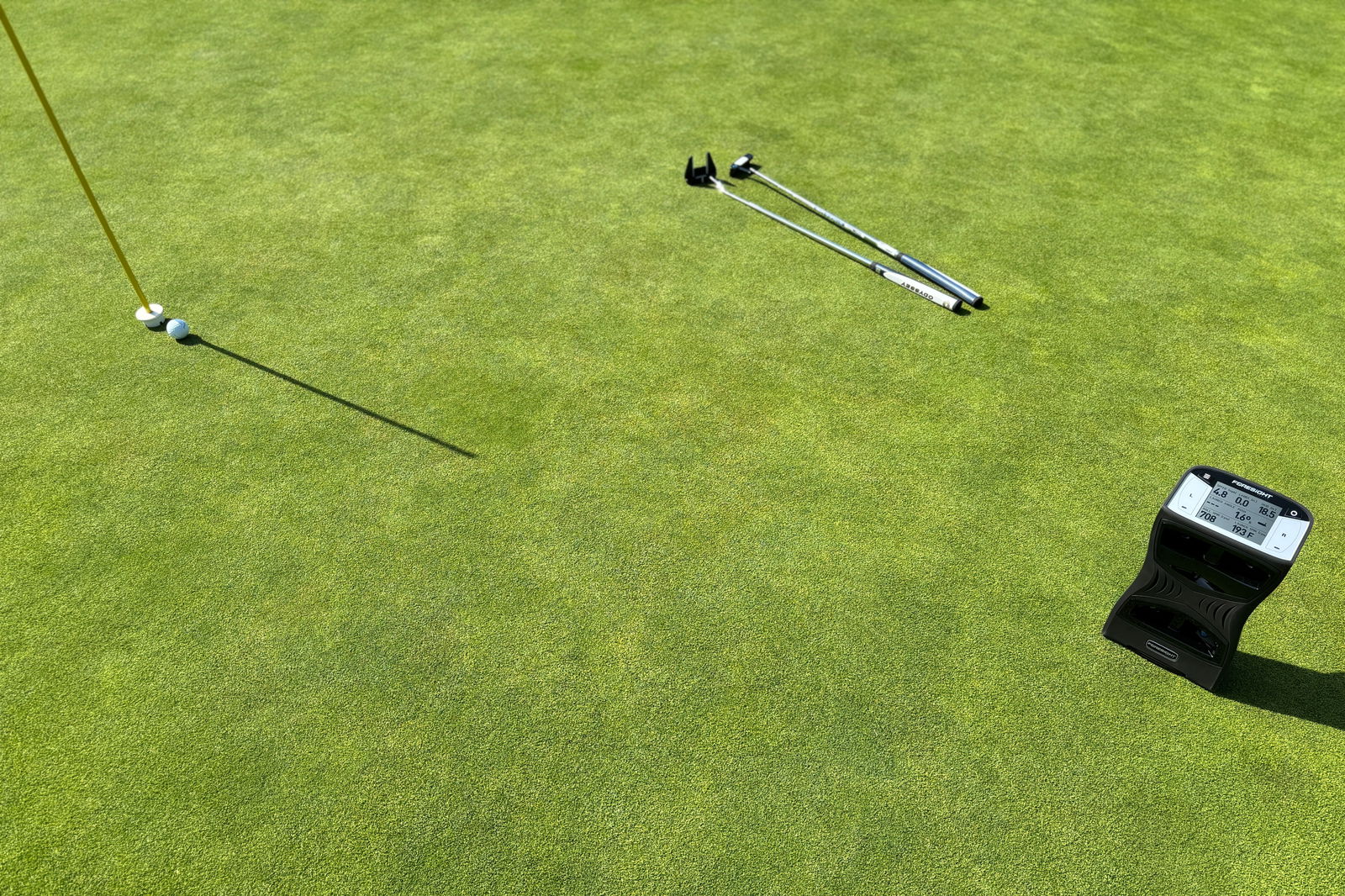 A Foresight Sports QuadMAX portable golf launch monitor sitting on the green near a flag with two putters