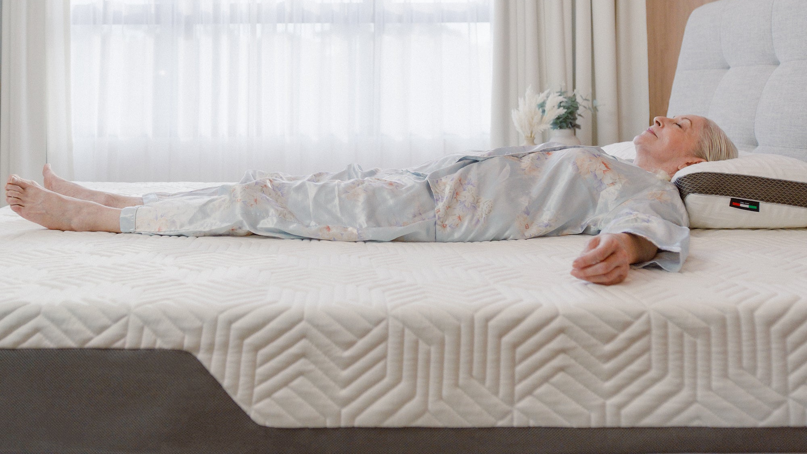 Valmori Hybrid II mattress lifestyle image depicting older woman in blue and floral pyjamas lying on top of white and grey mattress